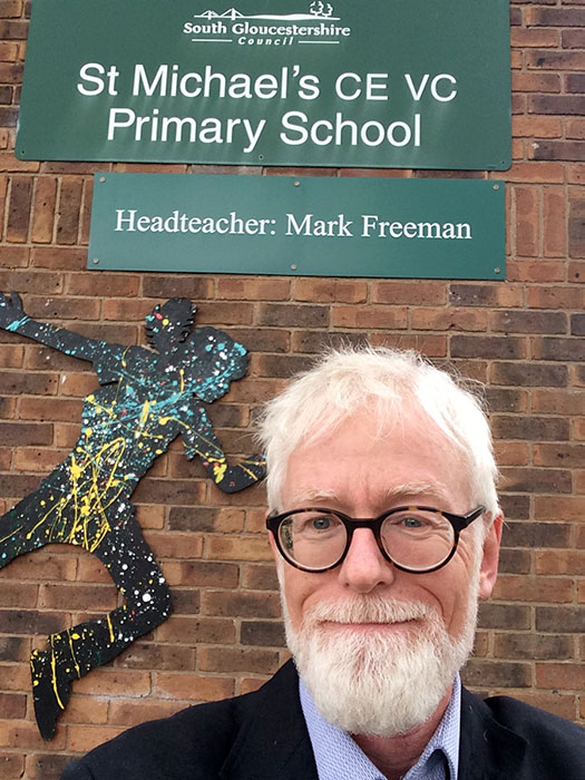 Photo of a man, with a school sign behind him.