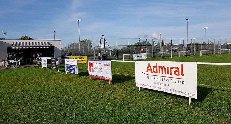 Photo of advertising boards on railings around a football pitch.