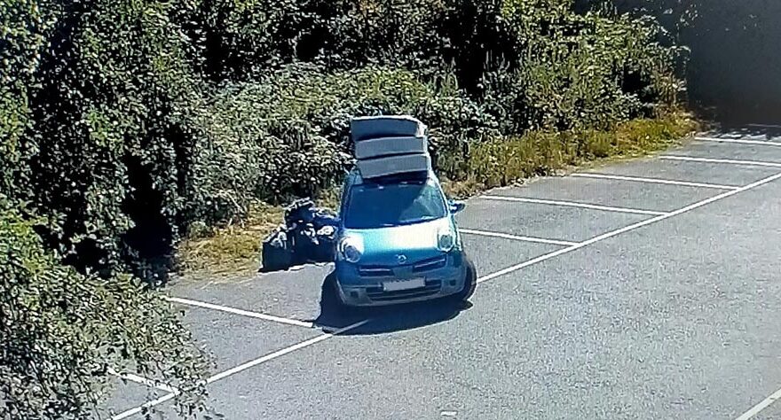 Photo of a car with mattresses on a roof rack and a number of black bin backs on the ground alongside it.